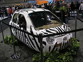 An extremely compact white car with black, zebra-like stripes behind black cordons