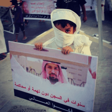 A sign board that has The Picture of Abdulla Majed Al Noaimi rising another board in one of his Human Rights protests. the sign board is held By a young brave eight years old kid.