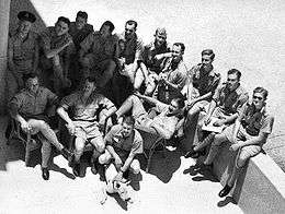 Informal high-angled portrait of fourteen men in light-coloured military shorts and shirts, with a dog in the foreground
