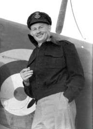 Informal portrait of grinning man in peaked cap and military uniform leaning against an aircraft fuselage