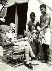 Caucasian man in light-coloured military uniform seated outside tent and being served tea by two indigenous men in light-coloured shorts and loincloth