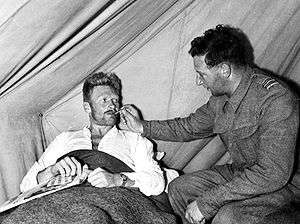 Blond man in bed having his temperature taken by another man wearing a military uniform