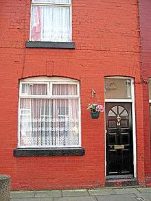 Exterior of a red brick building. Visible are a black door with a small window just above it, two larger windows to the left of the door, one above the other, and a flowerpot between the door and the lower larger window