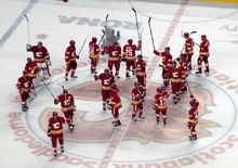Twenty hockey players in red uniforms stand at centre ice with their sticks raised in salute to the crowd around them.
