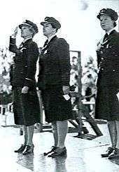 Three women in dark military uniforms standing to attention on a platform