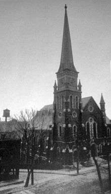 An ornate dark stone or brick church with a tall steeple seen in a black and white image from a three-quarter angle
