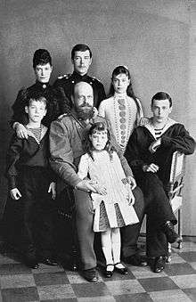 Black-and-white photograph of the Romanov family. Olga is a young girl who stands at the front resting against the arms of her seated bearded and bald father, who wears a military uniform. The older children and the empress complete the group.