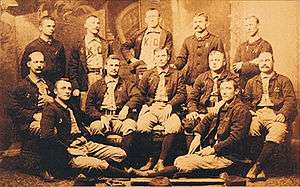 Baseball players are posing for a photograph, five men standing, five men sitting on chairs, and two are sitting on the floor.