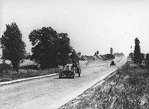 An open-topped sports car, with '3A' written on the front, kicks up dust as it drives along a road lined with trees and grass. Another open-topped sports car trails behind.