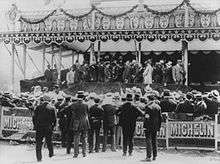 A crowd gathers in front of a canopied grandstand to watch a presentation ceremony. The bottom of the grandstand carries sponsorship by Michelin.
