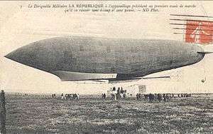 Postcard with orange stamp (French) affixed shows left flank of an airship just above the ground; some people are on the ground below the airship