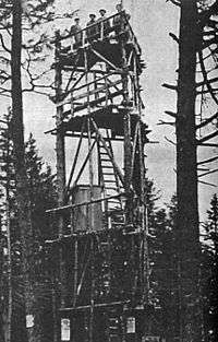A black and white photograph of a triangular wooden tower with an open top deck, with some evergreen trees in the foreground. Several men are standing on top of it.