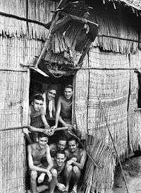 Shirtless and malnourished men smiling in a in the doorway of a hut