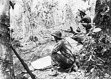 Black and white photo of a man wearing military uniform armed with a large gun lying down and aiming the weapon into dense bushland. Two other men in military uniform are crouched on either side of the prone man.