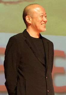 A male composer wearing a brown suit with his hands crossed behind his back and a smile on his face.