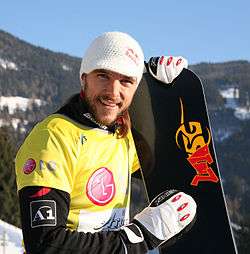 Man wearing a yellow snowboarding jersey and a white knitted cap. He is holding a black snowboard with gloved hands, and is standing on a snowy hill covered with trees.