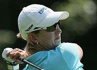 A blonde-haired woman in a white hat and light blue shirt holding a golf club at the end of a swing