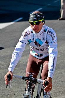 A road racing cyclist wearing a white jersey, stylised with blue and brown diamonds, and brown shorts.