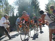 A line of three road racing cyclists, led by one in an orange jersey. Spectators line the road on either side, mere inches from the cyclists.