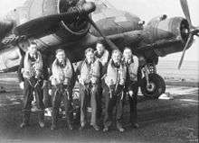 Six young men wearing flying suits standing in front of a World War II-era twin engined monoplane