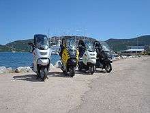 Four BMW C1 scooters parked side by side in front of a lake, with mountains and the masts of a large ship in the background
