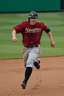 A white man in a red baseball uniform running bases. Looking into the camera, he has on a red baseball uniform reading  "Houston" in yellow text with similarly-colored line under it. Below that to the runner's left hand side, there is a black "2" with a red outline. In addition to a black baseball with an orange star, he has on gray pants, black socks, and black cleats.