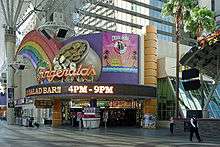 Ground-level view of the front entrance to the casino when it was called Fitzgeralds; a very prominent, colorful sign features a rainbow and a pot-of-gold. There are slot machines inside the building, and glass windows and part of a skyscraper are visible above the sign.