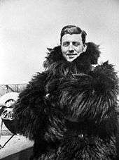 Half-length informal portrait of man in thick fur coat with a biplane in the background