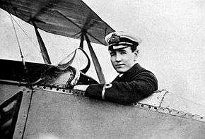 A man in military uniform with peaked cap, sitting in the cockpit of a biplane