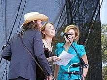 A man and two women crowded around a microphone and singing.  The man is on the left and wearing a dark blue suit and a white cowboy hat. Welch, in the middle, is wearing a black dress, and the woman on the right is wearing a green dress.
