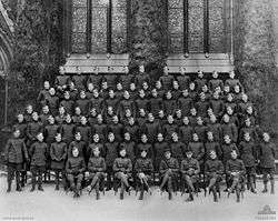 A large group portrait of men in military uniform. The men in the front row are seated, while six successive rows are standing behind them on elevated platforms. The men are standing in front of a large building with stained glass windows.