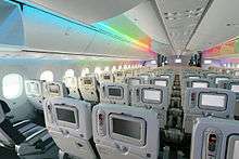 787 cabin. It shows the 787's spacious cabin. Above the blue seats are overhead bins and a rainbow light effect.
