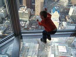young child leaning against and looking out a skyscraper window on a floor that is also glass