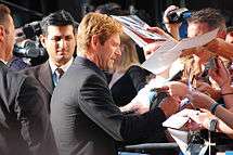 A caucasian male is signing autographs for fans. He has blond hair, and is wearing a black suit jacket. Visible in the background are other people.