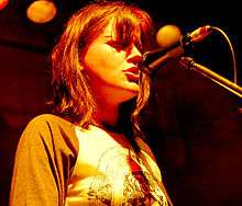 May is singing into a microphone on its stand. Her should length hair is brown and the fringe covers her eyes. She stares forward, wearing a brown sleeved tee-shirt with a design printed on front. Stage lights are visible behind and above her.