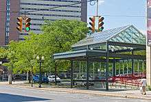 An urban street corner with a small area planted with small trees bordering a parking lot. An elaborate steel and glass structure shelters a wheelchair ramp going into a building at right. In the foreground are two suspended traffic lights, showing green; in the background is a high-rise