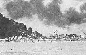 Black and white photograph depicting an aircraft on fire. The tail of another aircraft is visible at the right of the photo and there is a large amount of smoke in the air.