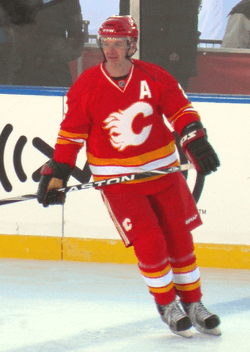 An ice hockey player looks to his right as he glides across the ice. He is in a red uniform with white and yellow trim, with a stylized "C" logo on his chest.
