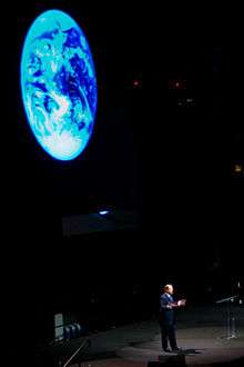 A man is standing on a stage in front of a giant projection of the Earth