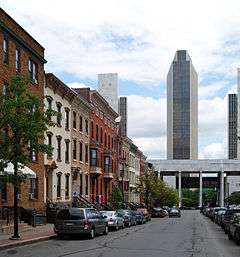 A view down a street shows 19th century row houses on the left, cars parked along the street, and tall, modern towers at the end of the street.