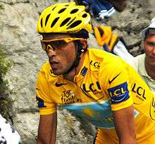 Close-up of a cyclist in a yellow jersey, wearing yellow-rimmed sunglasses and a yellow helmet. His bicycle is not visible, but he is in riding position. A spectator is partly visible behind him.
