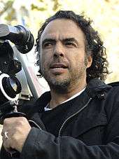 A man with long hair wearing a black coat with a camera in hand.