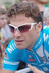 A man in his early thirties, wearing white-framed sunglasses with red lenses and a blue cycling jersey with white trim.