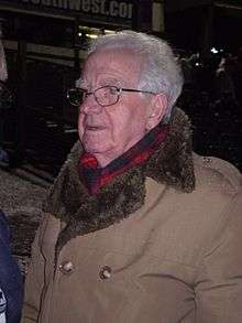 An elderly white man with grey hair and glasses wearing a warm coat and scarf, pictured outdoors at night.