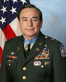 A color image showing Rascon from the waist up in his military dress uniform with ribbons. His Medal of Honor is visible around his neck and an American flag is visible in the background.