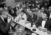 Malcolm X is holding a camera and taking a picture of Ali, who is sitting at a luncheonette counter