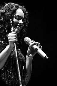 Black-and-white picture of a woman. She holds a microphone with her left hand. She wears a blouse with beads.