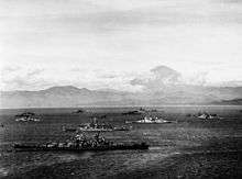 Black and white photo depicting 13 World War II-era warships anchored close together near the coast of a body of water. Steep mountains are visible in the background.