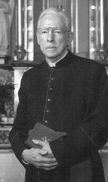 An older man dressed in Roman Catholic priestly garb holds two books in his hands, which he has folded in front of him, and stares at the camera with a neutral look on his face.