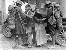 Two soldiers are warmly greeted by civilians – and elderly woman and man. A parked ambulance is behind them